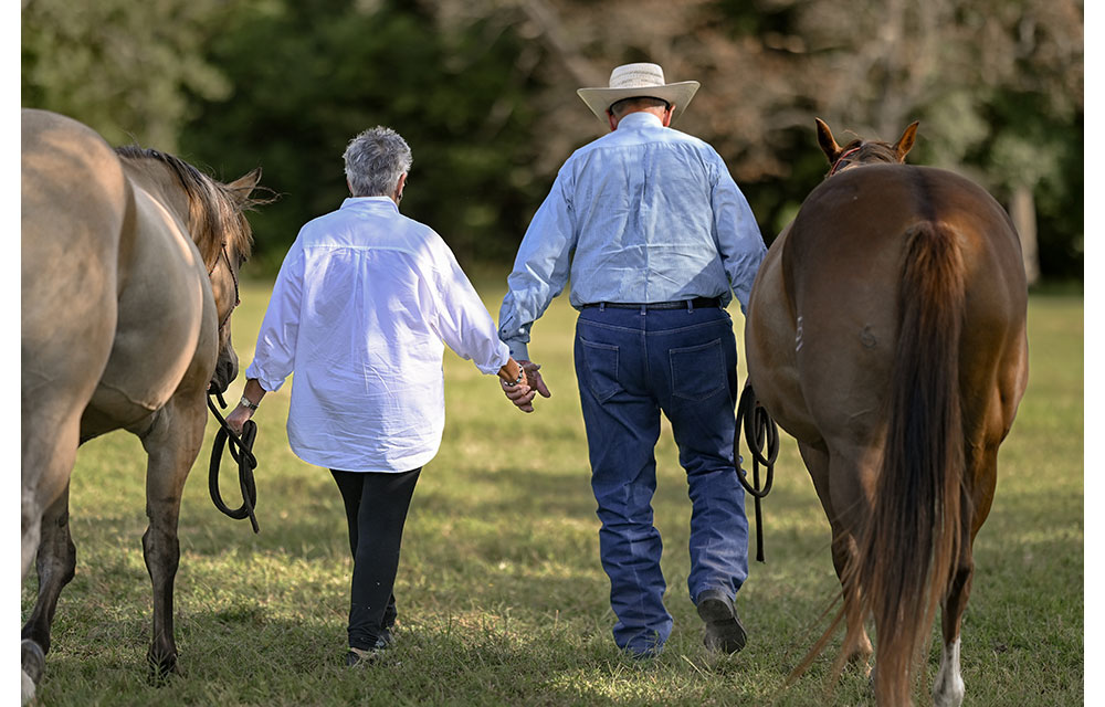 An older couple walks hand-in-hand through a pasture, leading two horses, with their backs turned to the camera.
